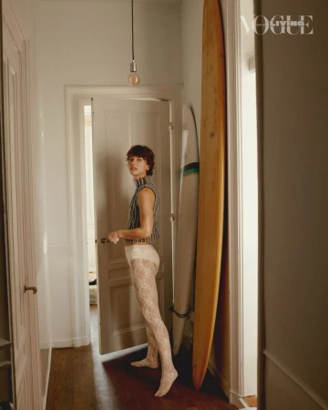Young adult woman in striped top and lace tights standing indoors near surfboards, styled for a Vogue editorial fashion shoot.