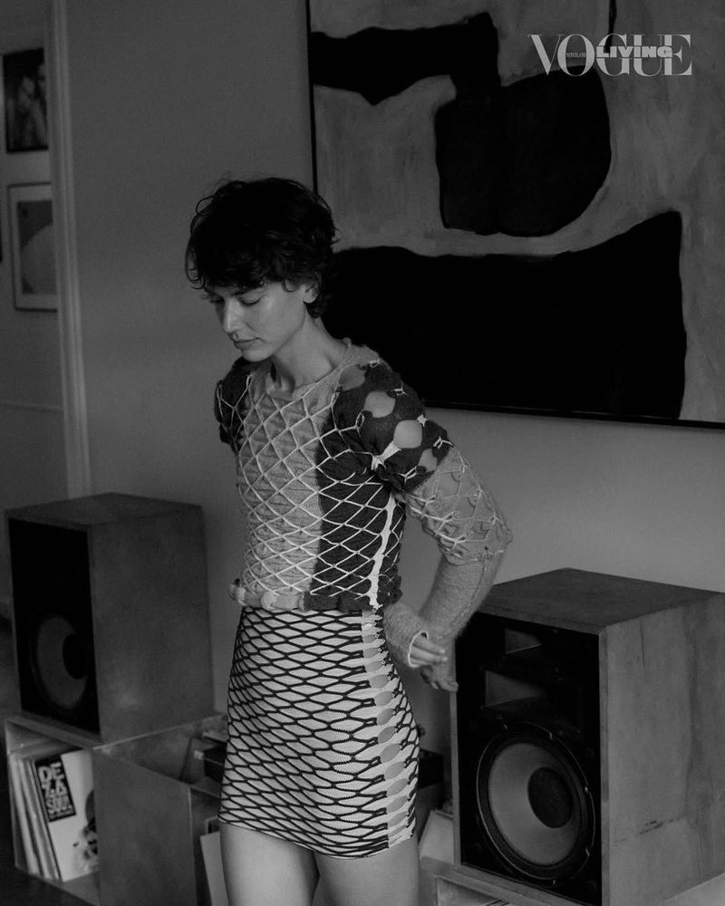 Black and white medium shot of a woman in a mesh top and skirt in an indoor setting, with speakers, art, and the Vogue logo visible.