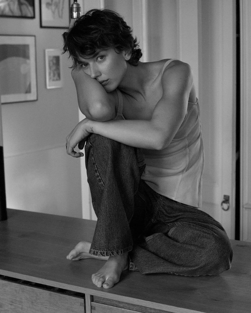 Monochrome portrait of a young woman in jeans and a tank top sitting barefoot on a table with framed pictures in the background.