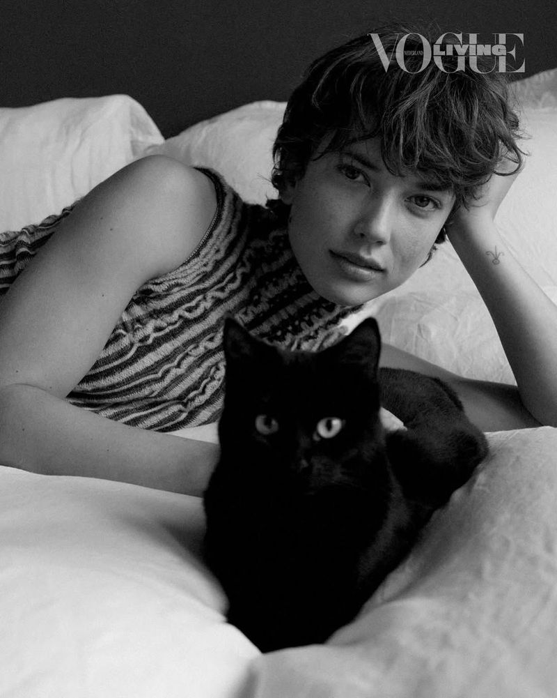 Black and white portrait of a woman with short hair and a black cat lying on a white bed under soft, natural light. Vogue Living logo visible.