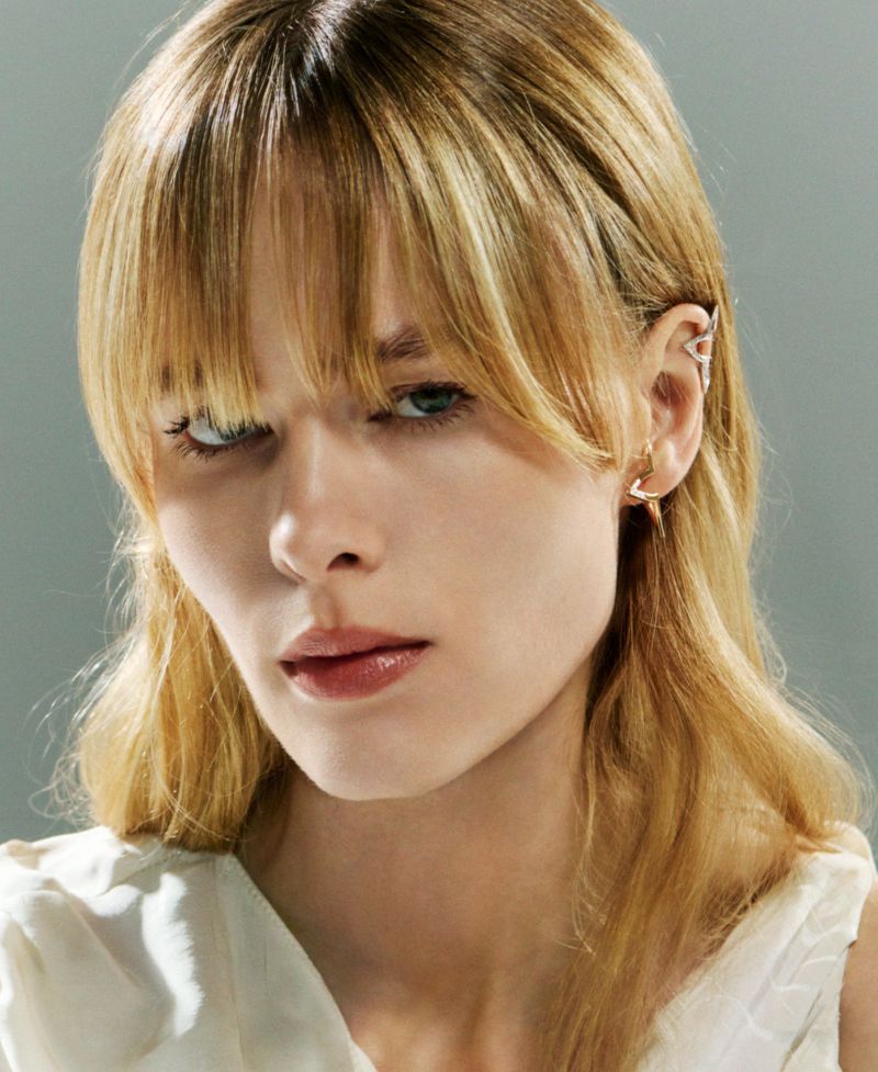 Close-up portrait of a blonde model wearing geometric silver earcuffs against a solid grey background.