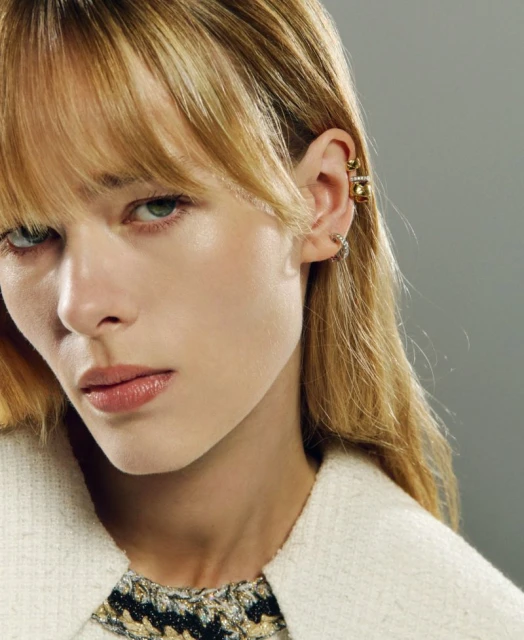 Close-up of a woman with blonde hair and fair skin wearing jewelry, including earrings and ear cuffs, posing against a gray backdrop.