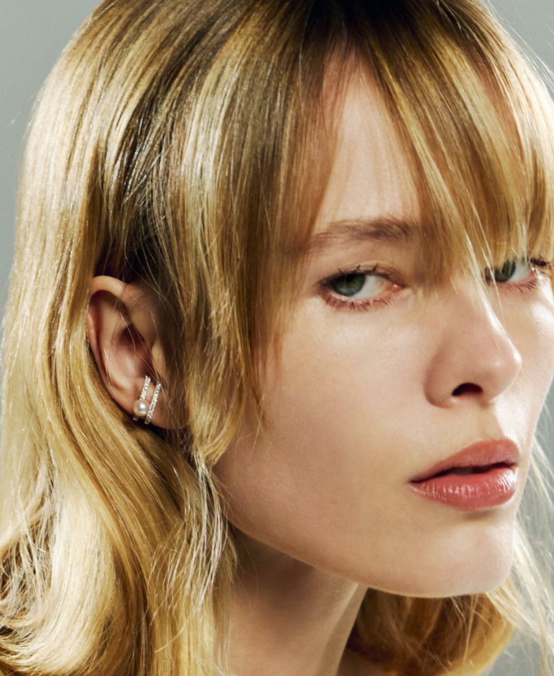 Close-up of a blonde woman with fair skin wearing silver earrings, posing indoors against a grey backdrop in a beauty setting.
