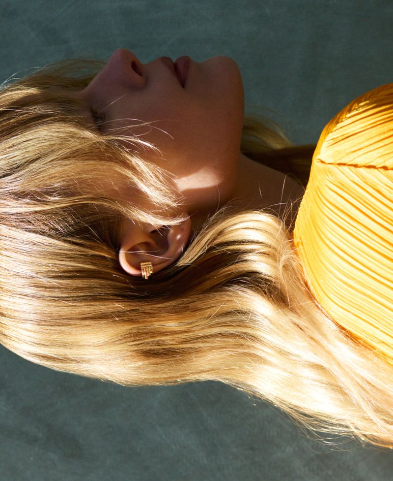 Close-up of an adult female with blonde hair, wearing a yellow top and small gold earrings, lying down against a textured light-blue surface.