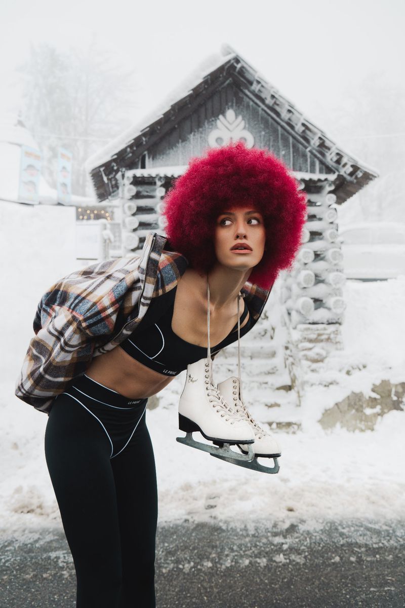 Woman with afro holds ice skates in snowy scene with cabin in background. She wears black activewear and a plaid jacket.