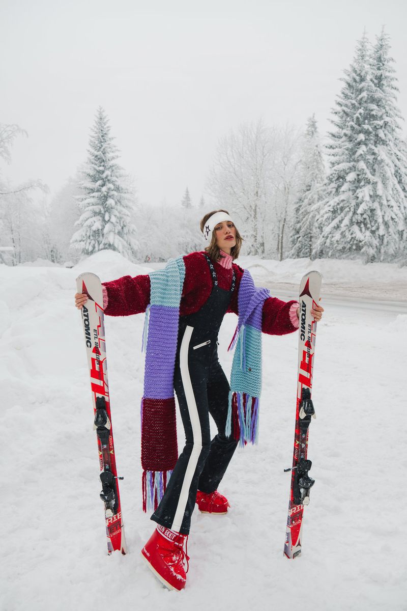 Woman in ski suit and red moon boots holds skis in a snowy mountain landscape setting with evergreen trees in the background.
