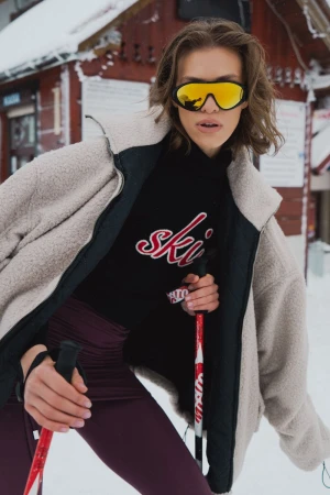 Adult female in ski goggles and outerwear holds ski poles in a snowy outdoor setting near a red building; outdoor fashion shoot.