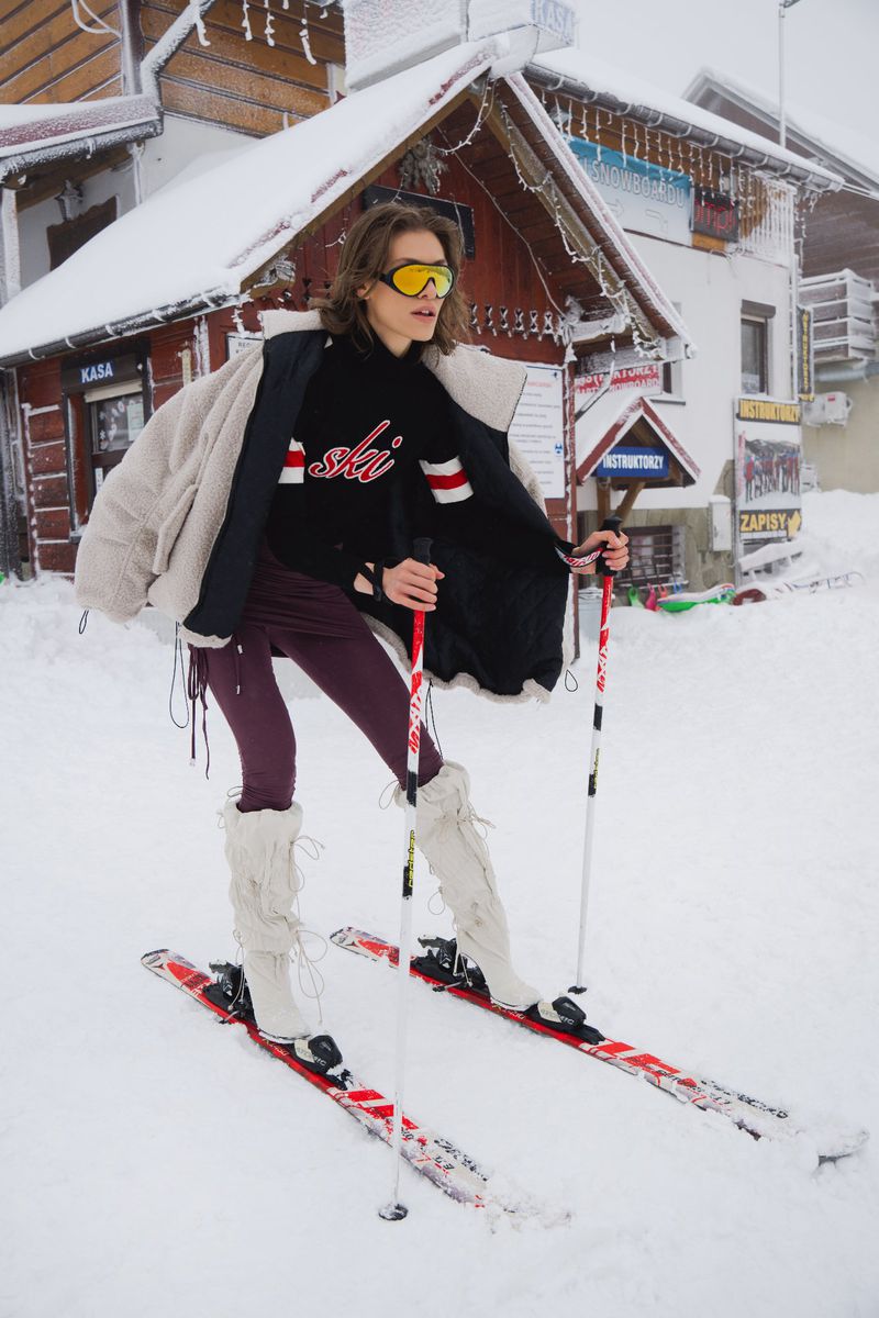 A woman in ski goggles poses in ski boots and skis in a snowy outdoor environment near a building with icicles and snowy rooftops.