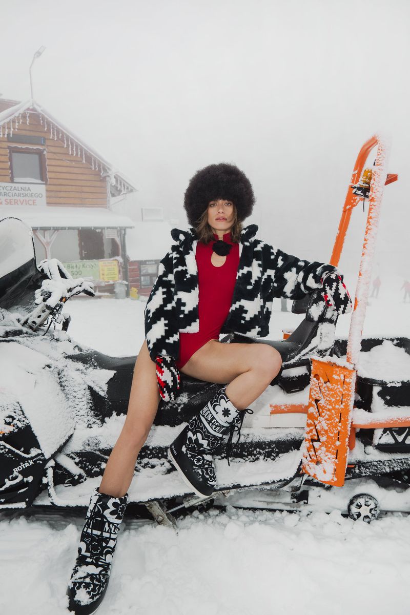 Woman in fur hat and patterned jacket sitting on a snowmobile in a snowy environment, wearing snow boots, for a winter fashion shoot.