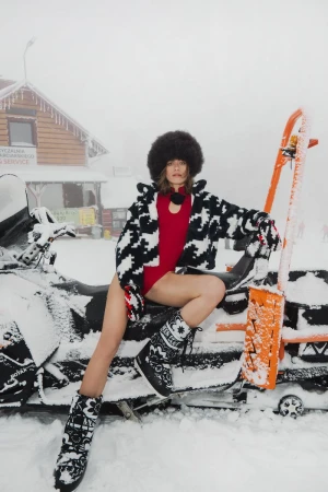 Woman in fur hat and patterned jacket sitting on a snowmobile in a snowy environment, wearing snow boots, for a winter fashion shoot.