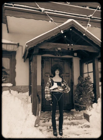 A person stands outside a snow-covered building while holding ice skates, wearing a patterned jacket, leggings, and a black top, with winter bulbs overhead.