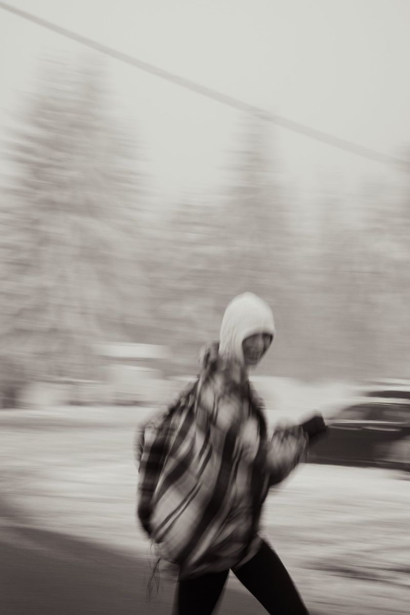 Person wearing a hooded jacket walks on a road. Background trees and car are blurred due to motion effect. Grey-scale tonality.