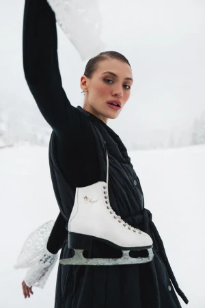 Elegant woman in a black coat with lace cuffs and a pair of white ice skates posing in a snowy, outdoor environment. Winter fashion.