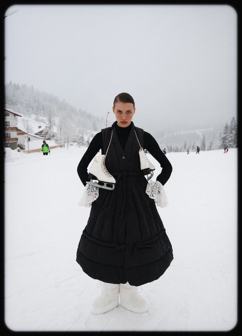 Woman in black dress on snowy hill carries ice skates. Winter landscape with trees and distant people. Outdoor fashion editorial.