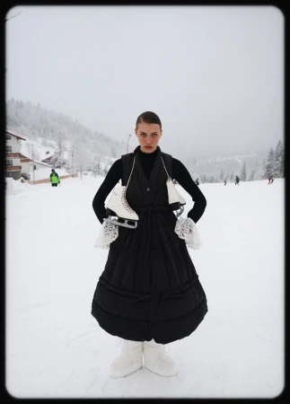 Woman in black dress on snowy hill carries ice skates. Winter landscape with trees and distant people. Outdoor fashion editorial.