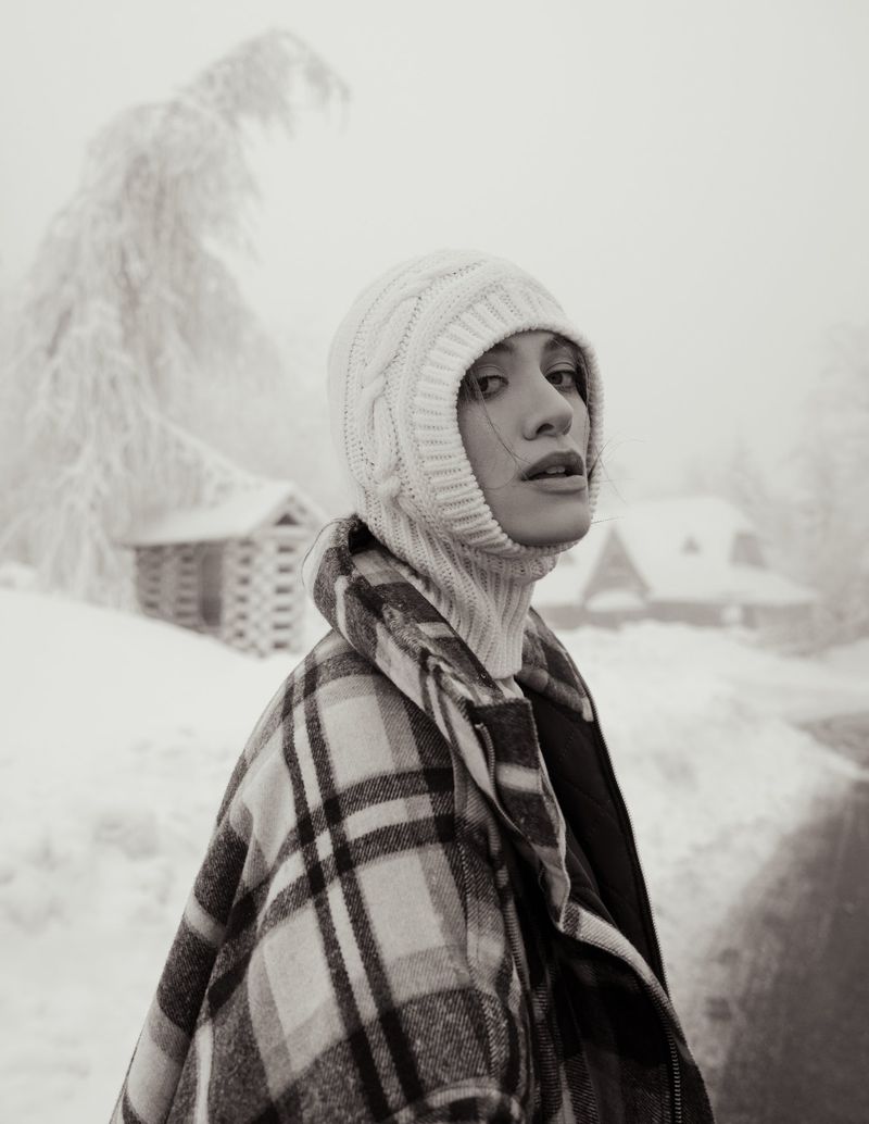 A woman in a white knit hat and plaid coat stands in a snowy landscape with cabin in the background in a subdued, black and white image.