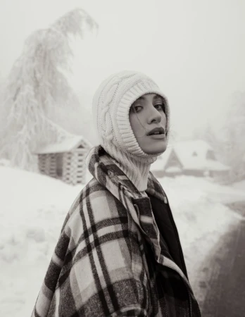 A woman in a white knit hat and plaid coat stands in a snowy landscape with cabin in the background in a subdued, black and white image.
