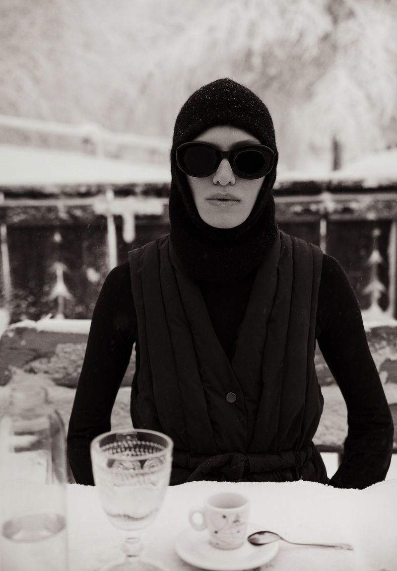 Woman in sunglasses and balaclava sitting at a snow-covered table with drinks and tableware in a wintry outdoor setting.
