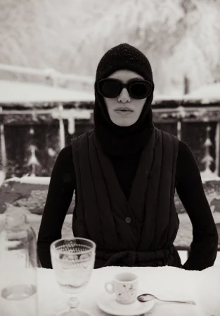 Woman in sunglasses and balaclava sitting at a snow-covered table with drinks and tableware in a wintry outdoor setting.