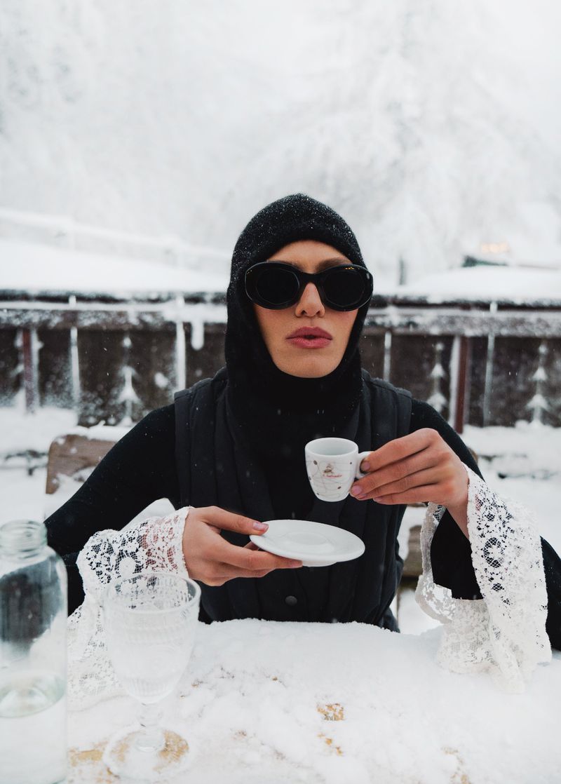 Adult in black balaclava, sunglasses sits at snow-covered table with coffee cup, saucer, and glasses against a blurred winter background.