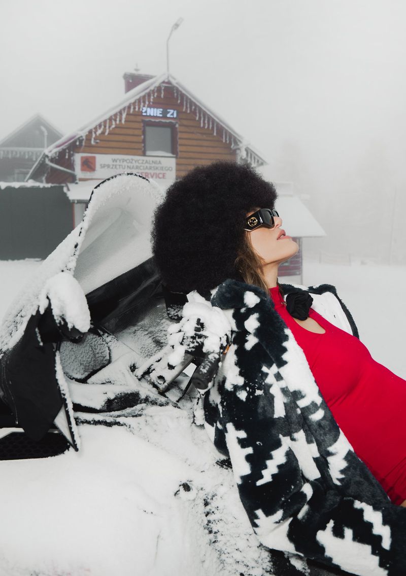 Woman in sunglasses and black fur hat poses outdoors near a snow-covered building on a foggy day wearing a red top and patterned jacket.