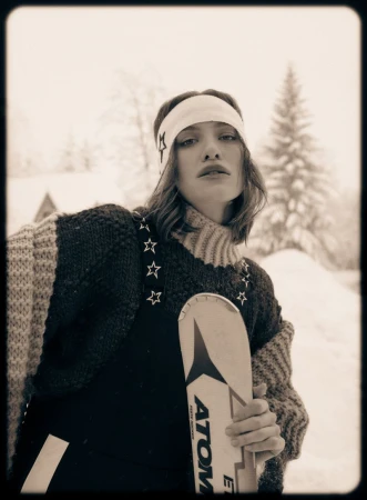 Portrait of a young woman wearing a star-patterned headband and sweater, holding skis in a snowy, rural winter landscape.