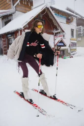 Model wearing yellow ski goggles, black sweater, and white boots on skis outside a wooden chalet in the snow