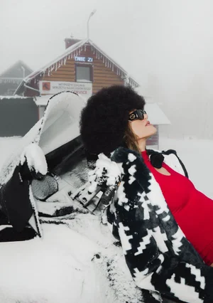 Model in black and white patterned coat and red top leaning against snowmobile in winter landscape