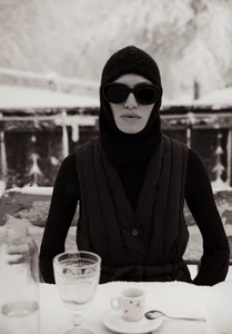 Model wearing dark sunglasses and a black hooded vest seated at a snowy table for L'Officiel Argentina Après ski editorial