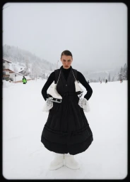 Model wearing a black quilted puffer vest and white snow boots holding ice skates in a snowy landscape