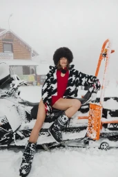 Woman in red bodysuit and black and white patterned jacket sitting on a snowmobile in a snowy mountain setting.