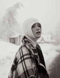 Model wearing a white cable-knit balaclava and plaid coat in a snowy winter landscape for an L'Officiel Argentina editorial.