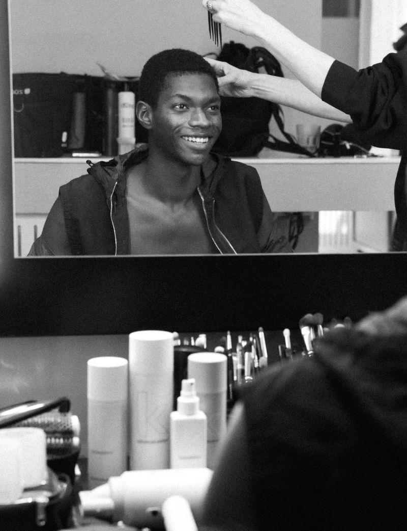 Reflection in a mirror shows a smiling Black male model having his hair styled in what appears to be a hair salon or photography studio.