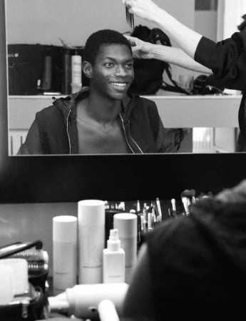 Reflection in a mirror shows a smiling Black male model having his hair styled in what appears to be a hair salon or photography studio.