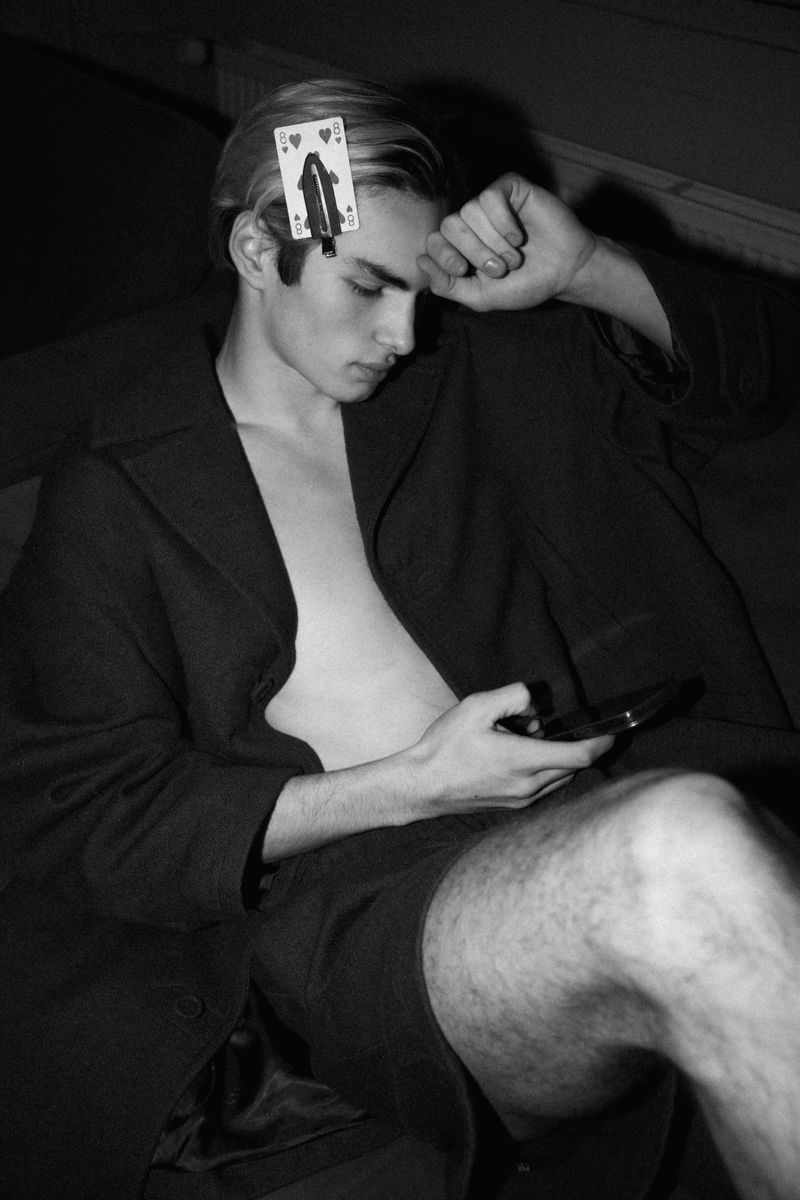 Monochrome portrait of a young man with a playing card on his forehead, holding a phone, wearing a coat, set indoors.