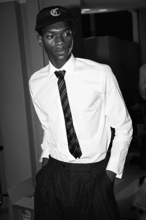 Black man wearing a white dress shirt, striped tie, cap, and dark trousers in an indoor setting.