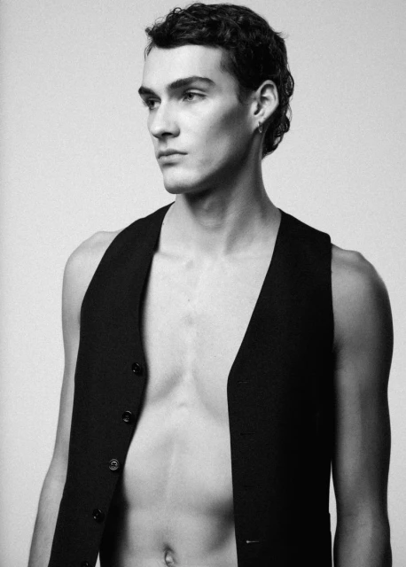 Black and white portrait of a young adult male with dark hair and a vest, looking off-camera against a neutral background.