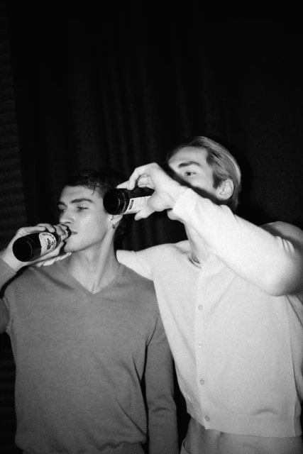 Two adult males in a black and white image, indoors, drinking bottled beverages with a dark curtain in the background, eye-level.