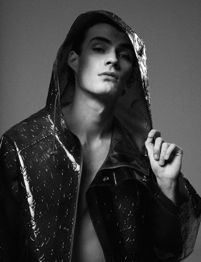 Monochrome portrait of a young adult male wearing a patterned, hooded jacket in a studio setting with a neutral gray background.