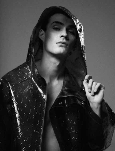 Monochrome portrait of a young adult male wearing a patterned, hooded jacket in a studio setting with a neutral gray background.