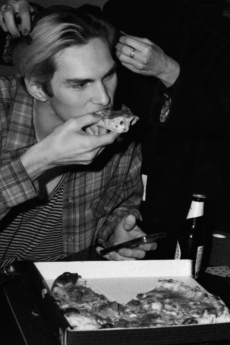 Indoor, medium shot shows a young adult eating pizza; a hand reaches into his blonde hair. Beer bottle, smartphone on the table.