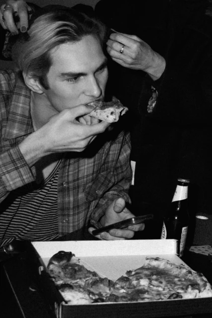 Indoor, medium shot shows a young adult eating pizza; a hand reaches into his blonde hair. Beer bottle, smartphone on the table.