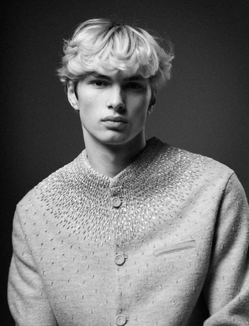 Black and white portrait of a young adult male with blond hair wearing a beaded jacket in a photo studio setting.
