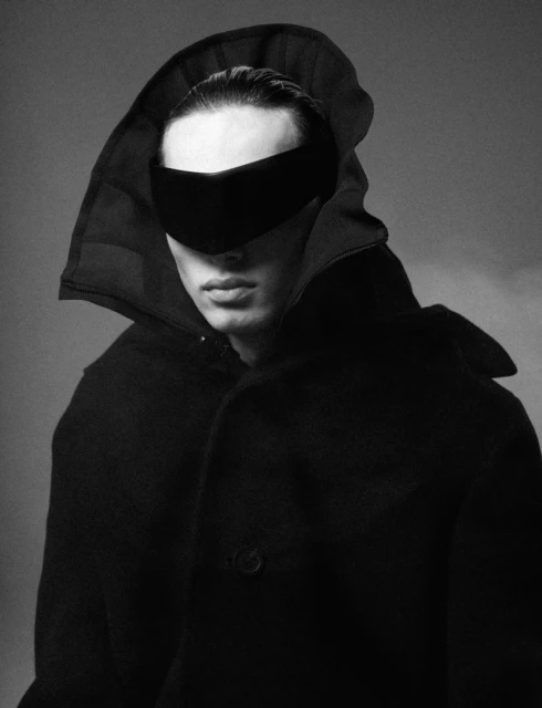 Portrait of a dark-haired male fashion model wearing a black hooded coat and black band over the eyes in a studio setting.