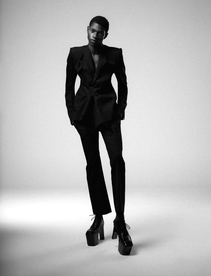 An androgynous Black male model in a black suit and platform shoes poses in a fashion photo studio with a white backdrop and soft lighting.