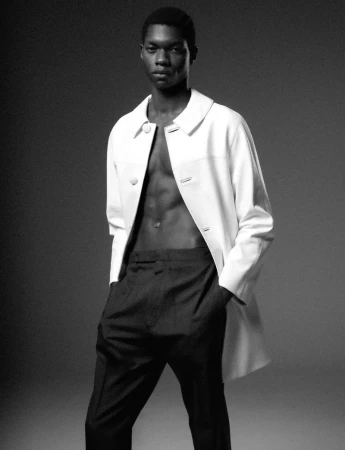 Black and white studio portrait of a male model wearing an open white jacket and dark trousers against a grey seamless background