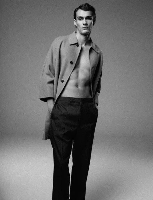 Studio shot of a male model in a light brown coat with no shirt, and black trousers against a grey backdrop, in a fashion pose.