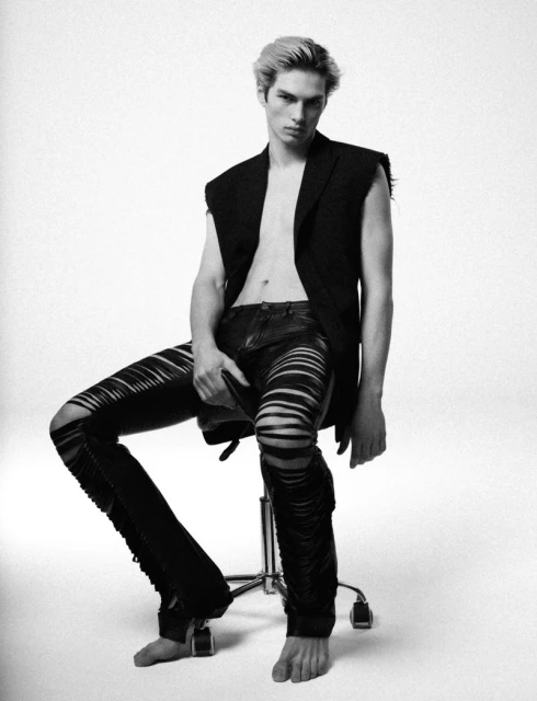 Young adult male model with blond hair poses in ripped leather pants and an open black vest while seated in a stark white studio.
