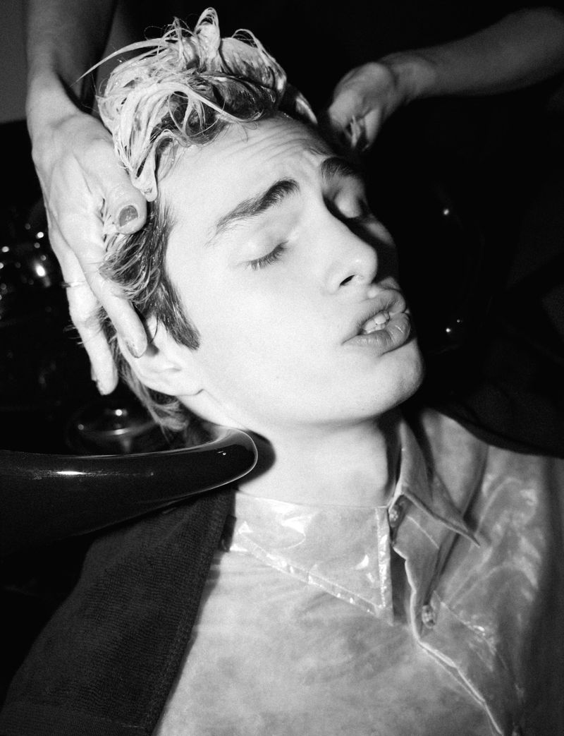A young adult male getting his hair washed in a black and white image, with hands massaging his scalp, and a shiny shirt.