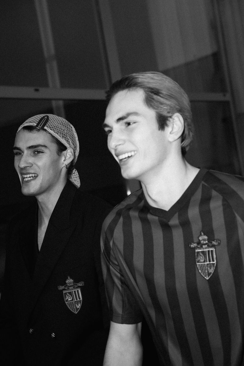 Two young adult men smile in a black and white indoor shot, wearing a jacket and striped shirt with crests, suggesting a vintage fashion vibe.